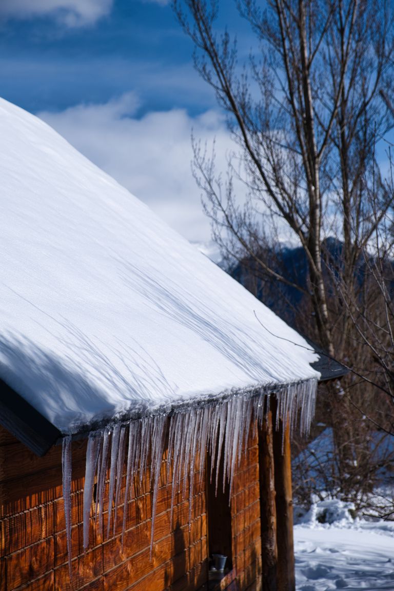 A snow-covered cabin with icicles under a clear blue sky, surrounded by bare trees.